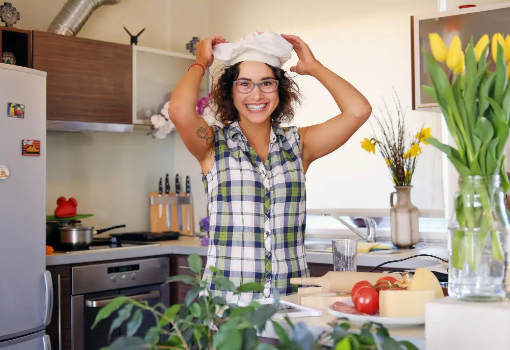 About 1 Sabrine smiling in her kitchen with a chef’s hat and fresh veggies.